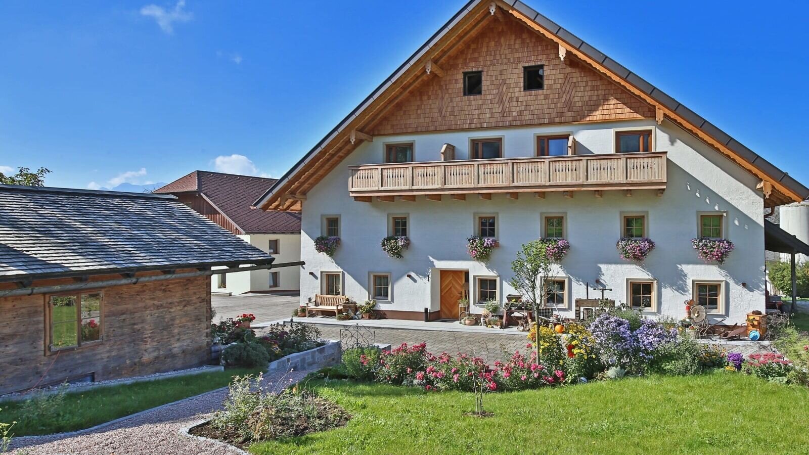 The main building of the farmhouse, featuring a wooden balcony, flower boxes, and a front garden with a lawn and gravel path.
