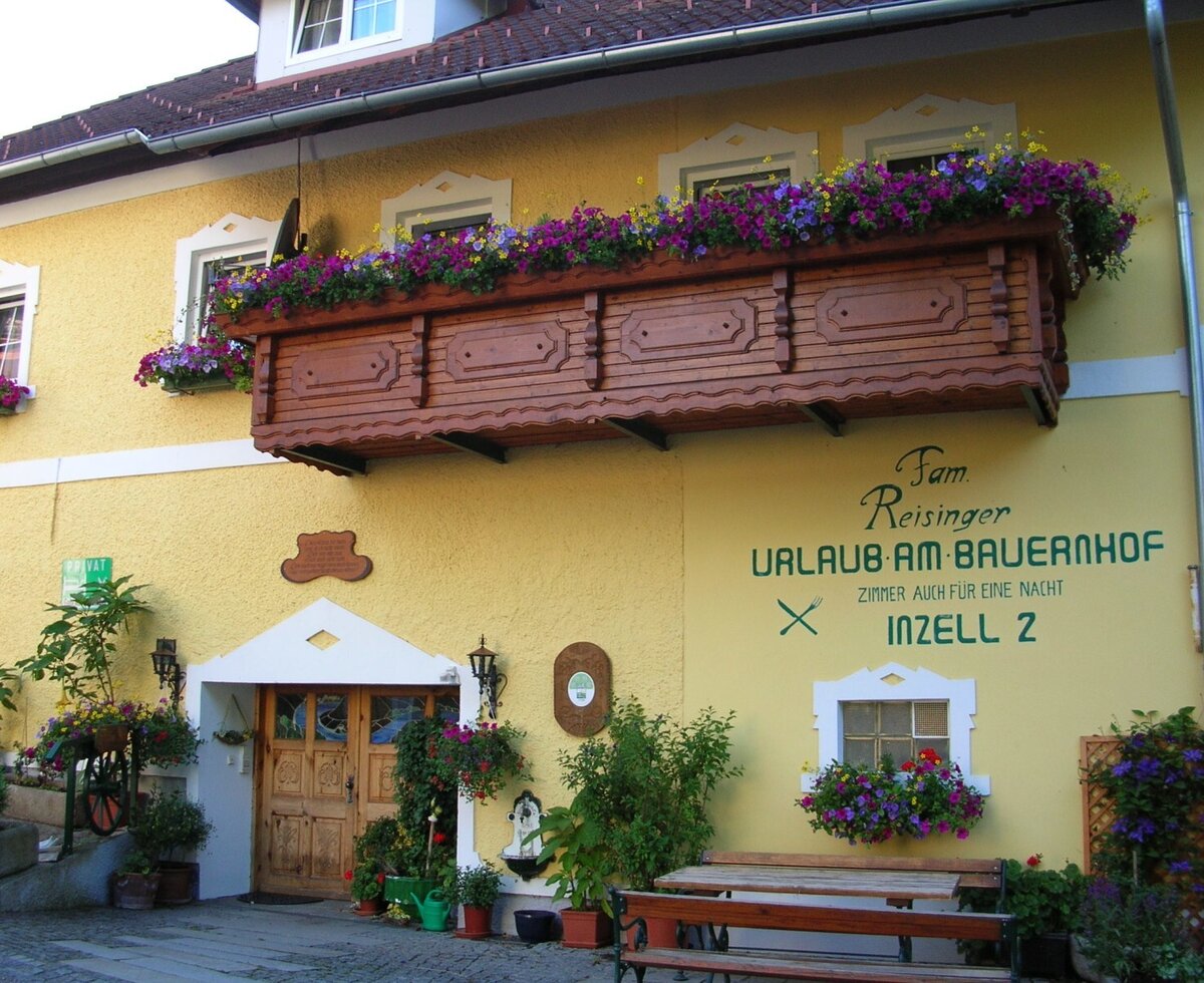 The Farm House exterior, featuring a yellow facade, a wooden balcony with flower boxes, and an outdoor bench and table.