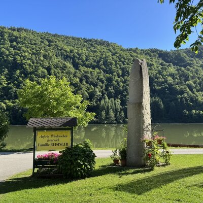 The outdoor area of the Bauernhof featuring a sign from the Reisinger family, a stone monument, and a view of the lake and forested mountains.
