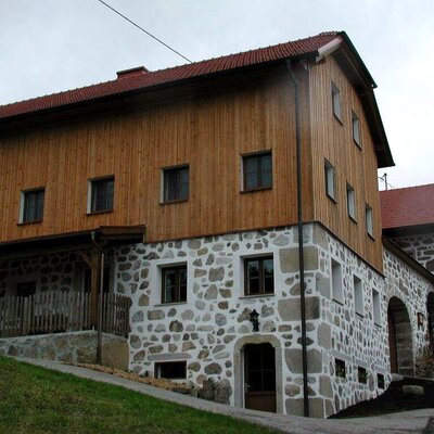 The exterior of the farmhouse, featuring a stone lower level, wooden upper levels, red tile roof, and a covered porch.