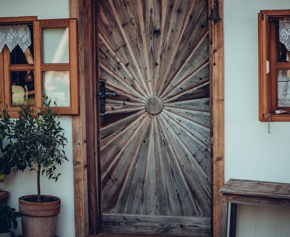 The main entrance of the farmhouse features a wooden door with a sunburst design, traditional wooden-framed windows with lace curtains, and potted lemon trees.