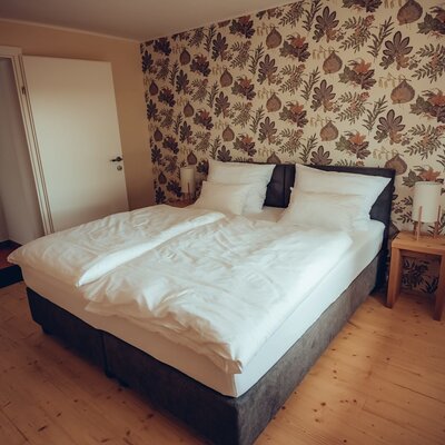 Double bedroom in the Farm House featuring a bed with white linen, a botanical-patterned accent wall, wooden flooring, and an open doorway.