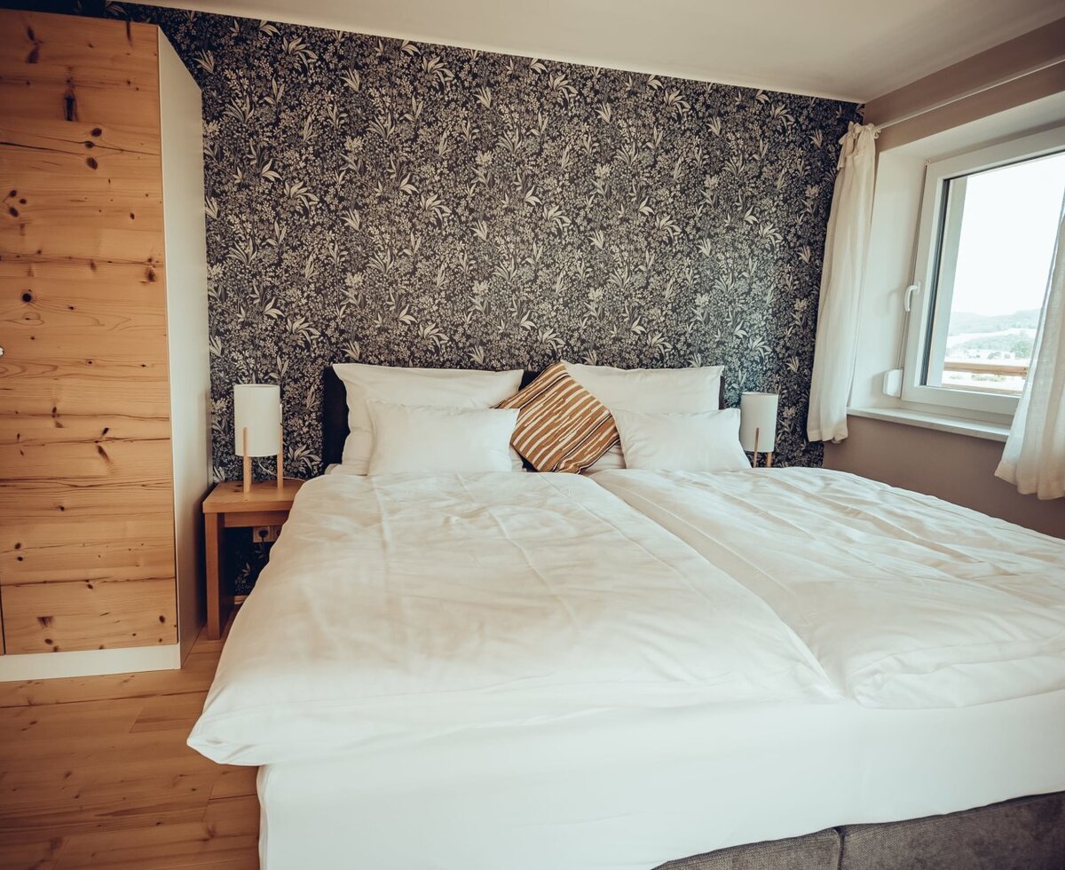 The Farm House bedroom features a double bed, patterned accent wall, wooden wardrobe, and a window with a view of the hills.