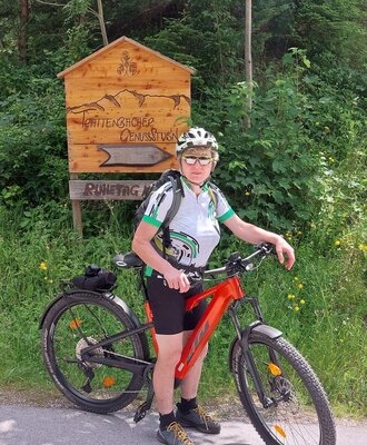 A cyclist with an e-bike in front of a signpost for Trattenbacher Genussstubn in the area surrounding the farmhouse.