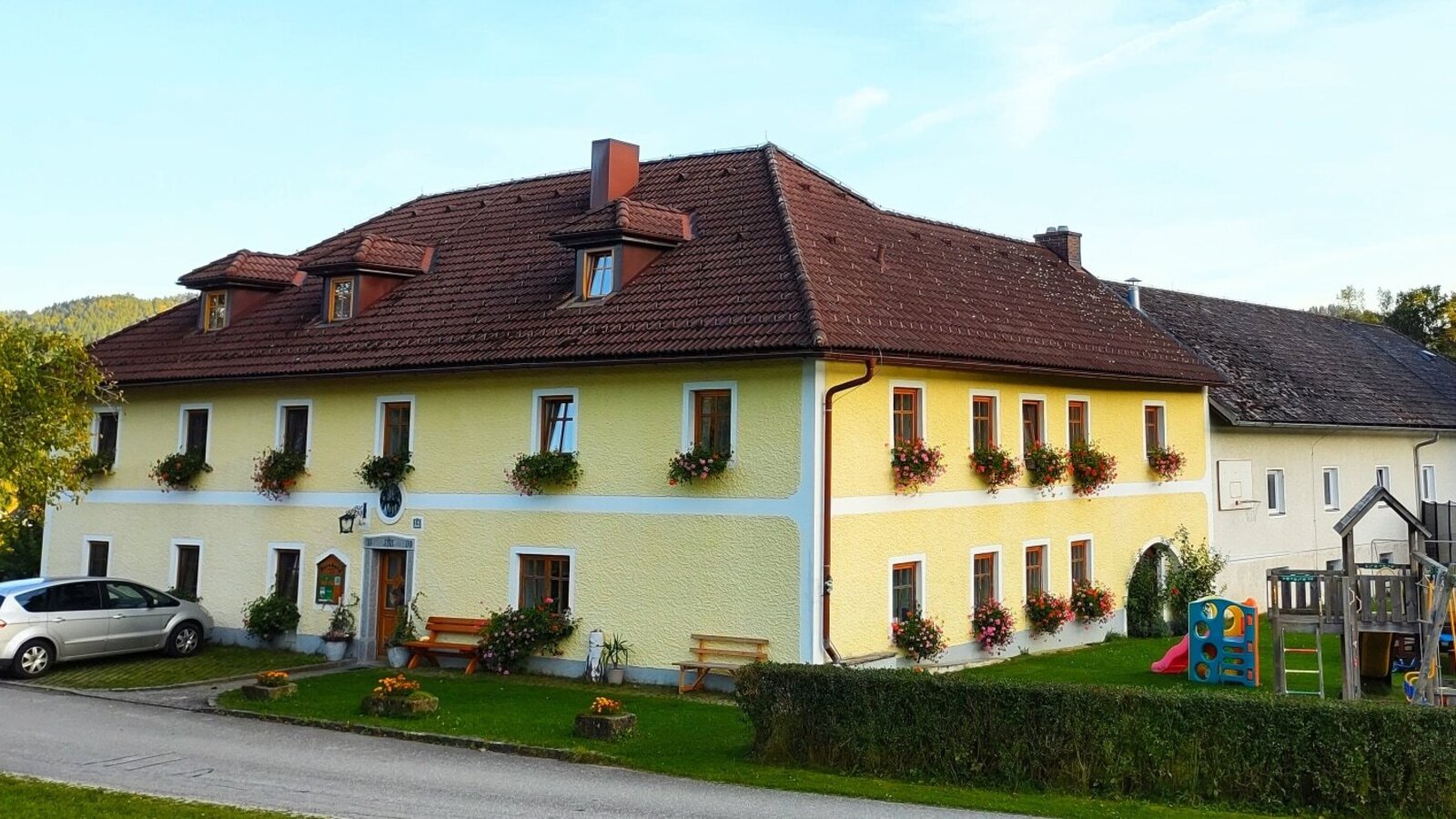 The yellow farm house exterior, with flower boxes on the windows, a parking area, and a children's playground on the lawn.