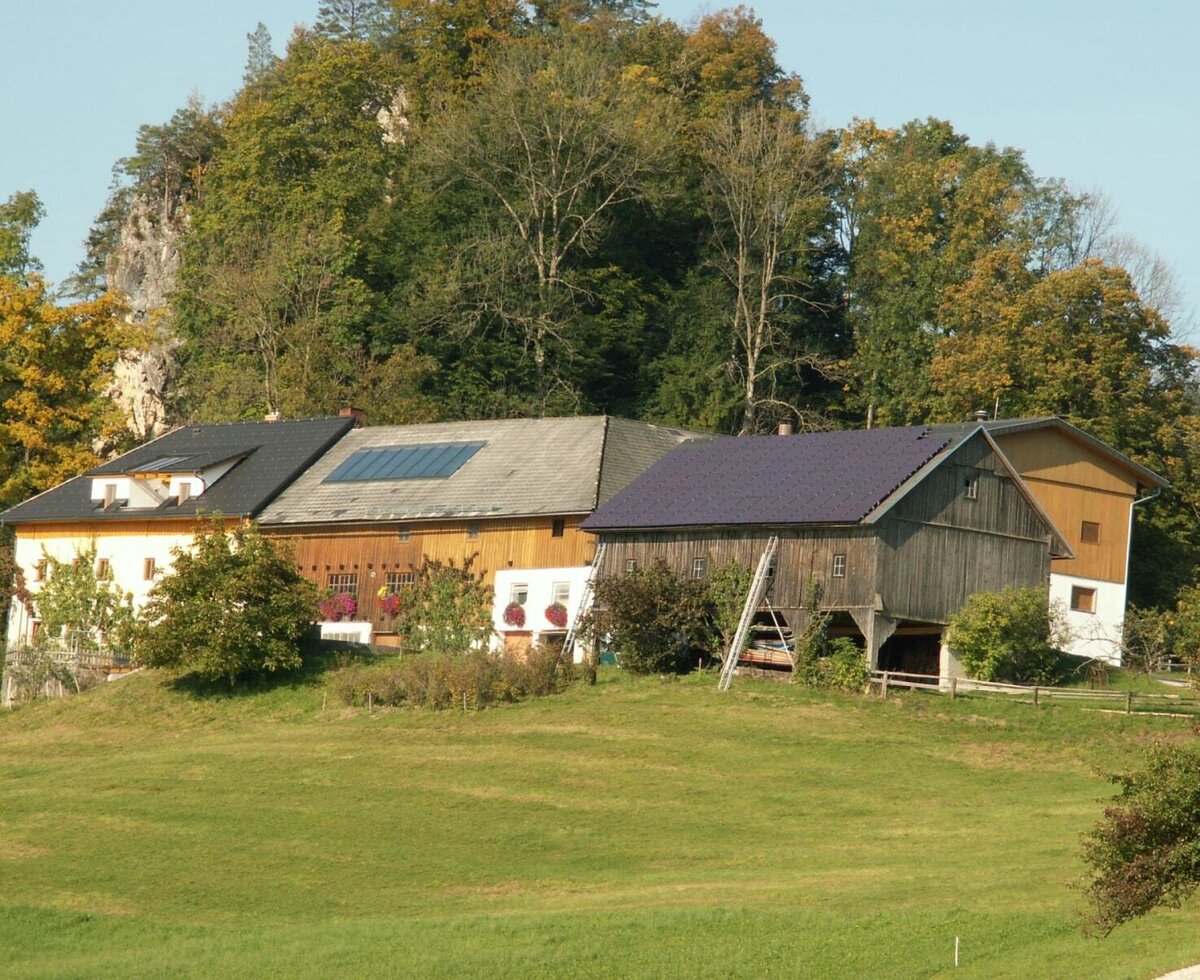 Exterior view of the farmhouse with a residential building, barn, and solar panels, surrounded by green meadows and trees.