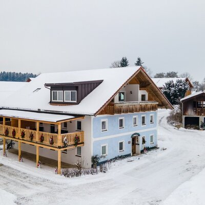 Exterior view of the Bauernhof in winter, featuring snow-covered roofs and a large wooden balcony on the light blue main building.