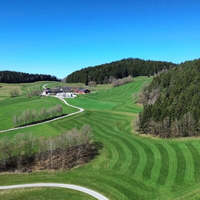The farmhouse is situated amidst green fields and wooded hills, with an access road.