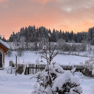 The winter landscape near the farmhouse, featuring a snow-covered chapel, forest, and an orange and pink sky.