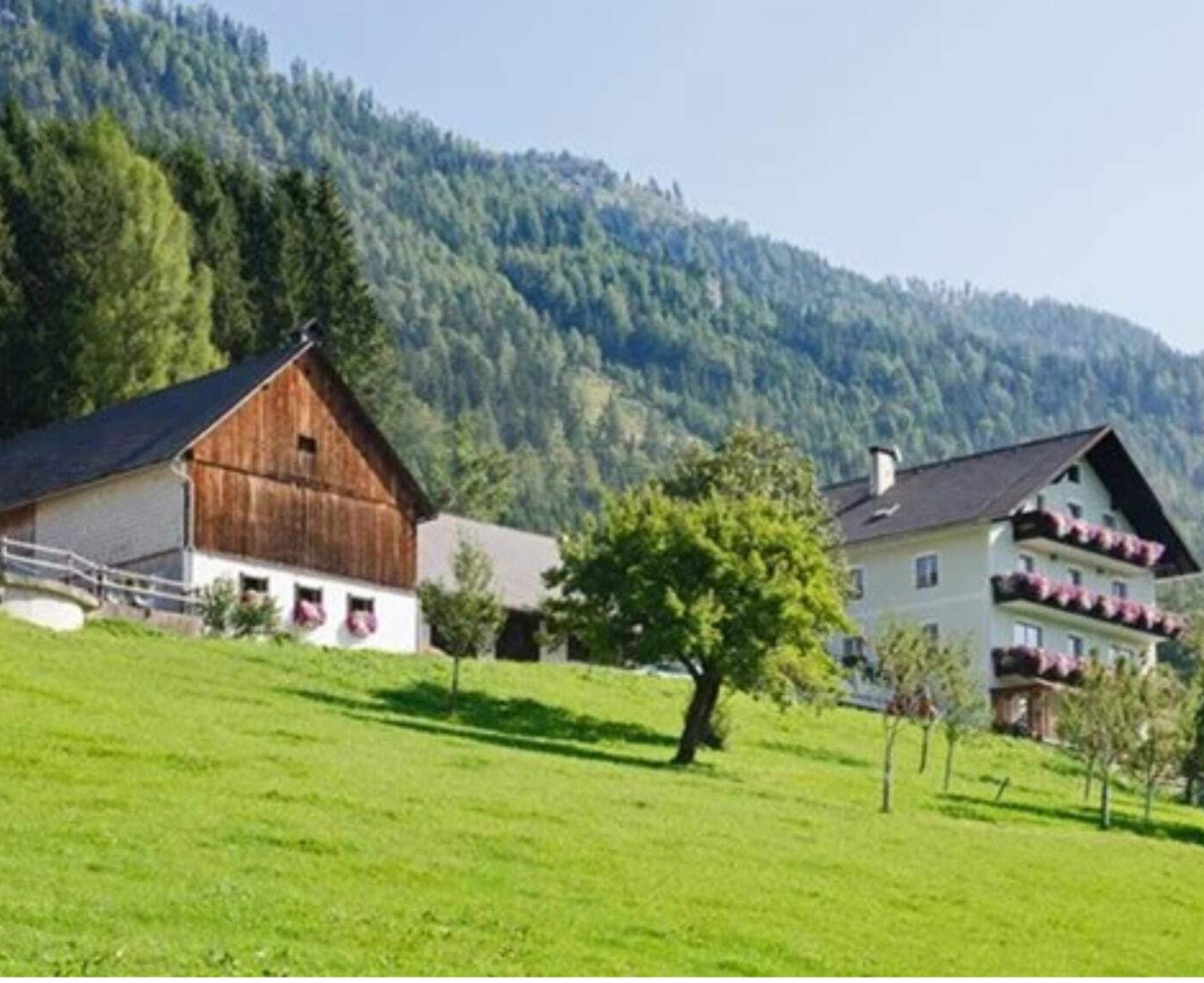 Exterior view of the farmhouse with a residential building with balconies and a farm building, surrounded by green hills and mountains.