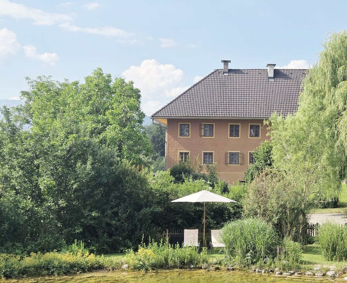 The idyllic exterior view of the farmhouse apartments, featuring a natural swimming pond, lush greenery, and relaxing sun loungers.