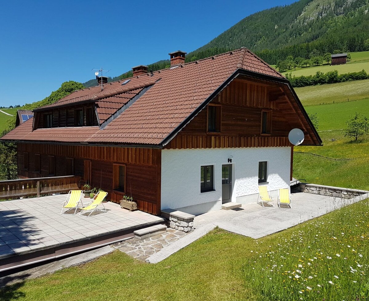 The exterior of the Farm House, featuring two paved terraces with sun loungers and a backdrop of green mountains.