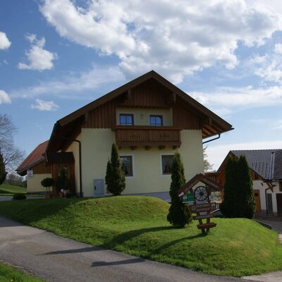 The farm house features a light yellow exterior with a wooden balcony and a well-kept lawn.