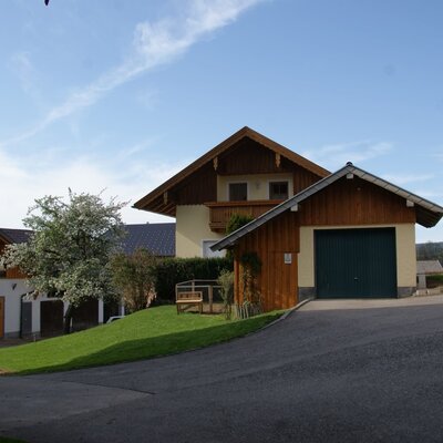 The Farm House exterior, featuring a wooden balcony, an attached garage, and a grassy lawn with a wooden bench.
