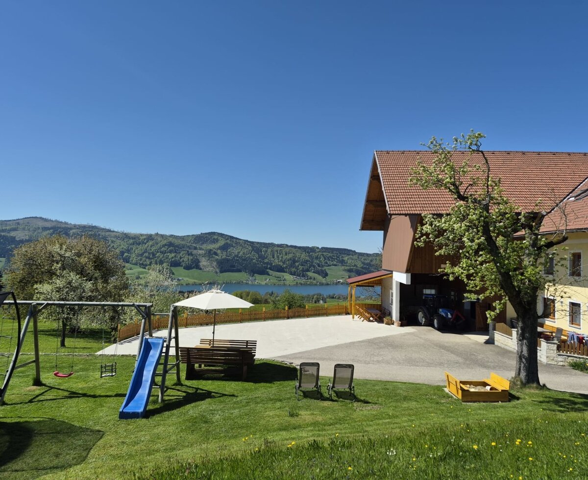Exterior view of the farmhouse with a garden, children's playground, seating, and a view of the lake and mountains.