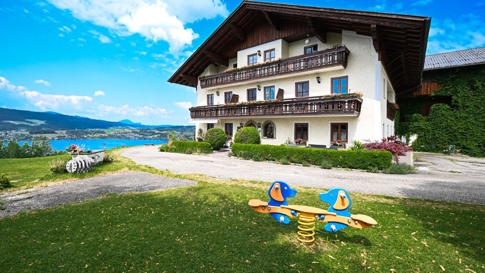 The farm house exterior features balconies, a children's spring rider on the lawn, and views of the lake and mountains.