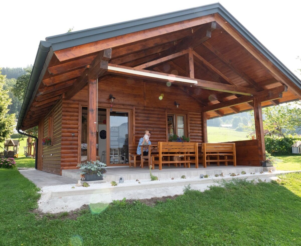 The wooden farm house features a covered patio with a wooden dining set, surrounded by a lawn and views of the surrounding hills, with a children's play area visible.
