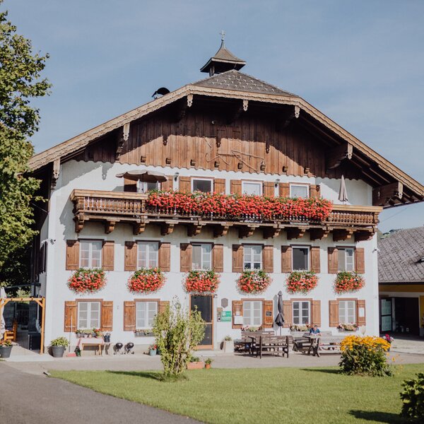 The farm house features balconies with red flowers, windows with wooden shutters, and an outdoor seating area with wooden tables and benches on a paved terrace.