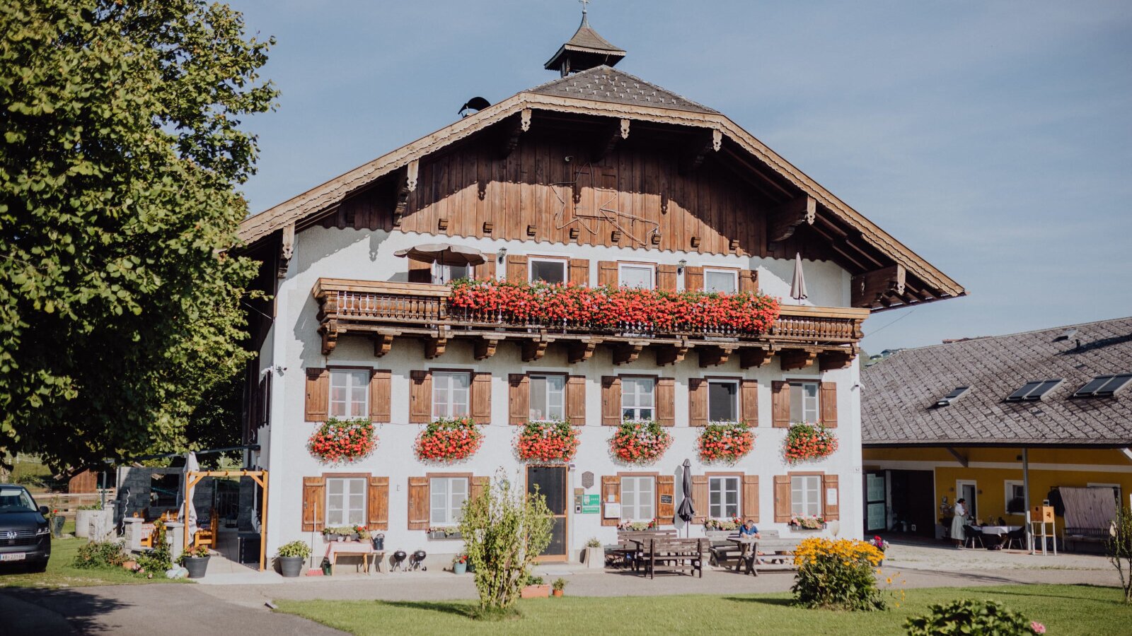 The farm house features balconies with red flowers, windows with wooden shutters, and an outdoor seating area with wooden tables and benches on a paved terrace.