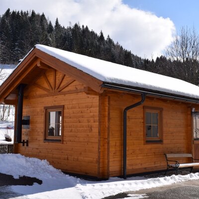 A garden house at the farm house, featuring a snow-covered roof, an exterior heating pipe, and a small bench by the door.