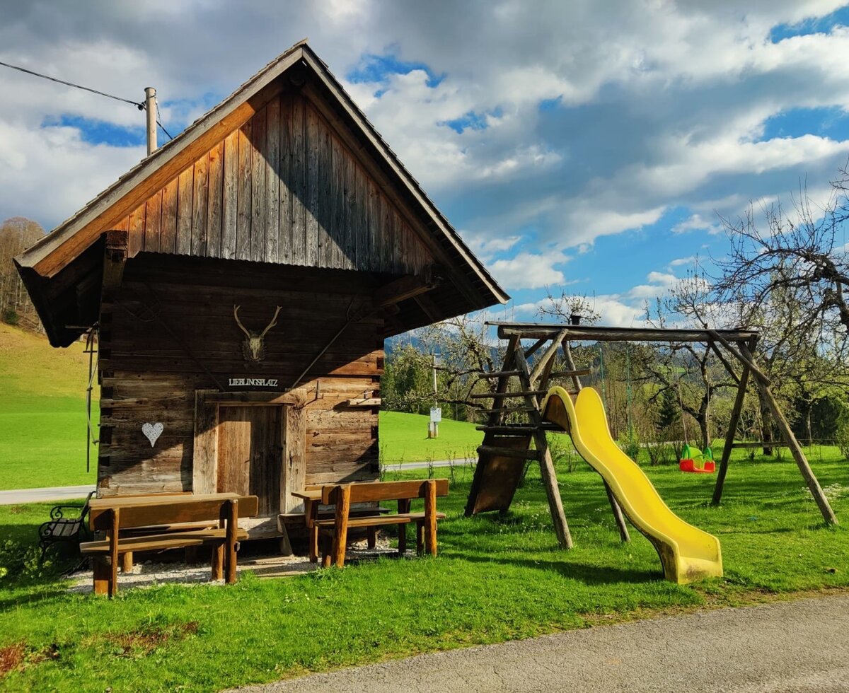 Playground with a slide and swings, and a wooden hut with seating at the farmhouse.