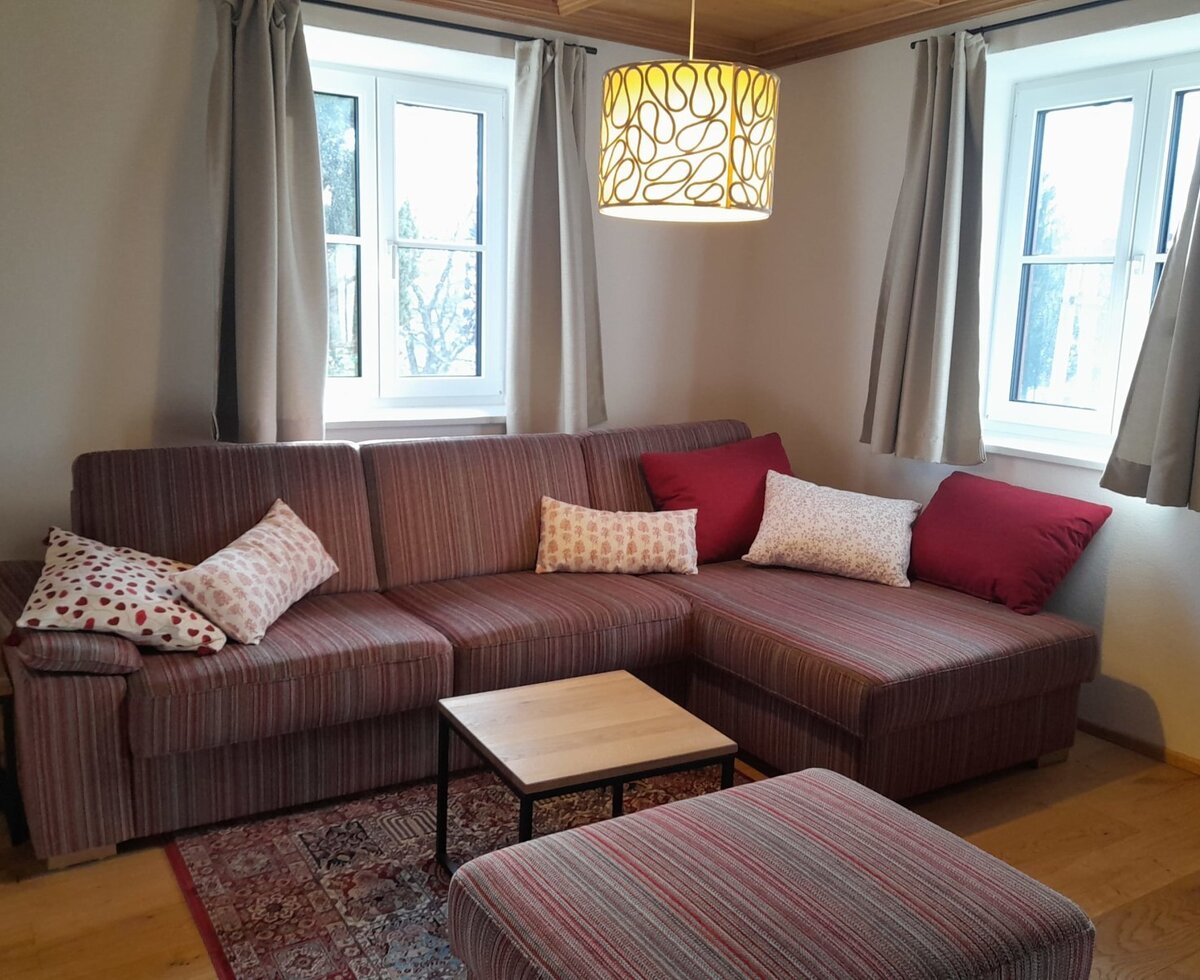 The living area of the farmhouse provides an L-shaped sofa with throw pillows, a coffee table, and an ottoman, illuminated by two windows.