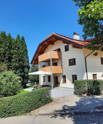 Exterior of the farmhouse, featuring wooden balconies, a tiled roof with solar panels, and an outdoor patio area with a picnic table and umbrella.