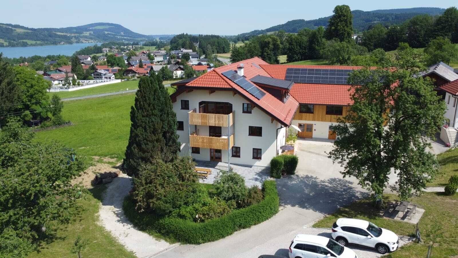 The Farm House features multiple wooden balconies, solar panels on its roof, an outdoor patio with seating, and offers views of the lake and village.