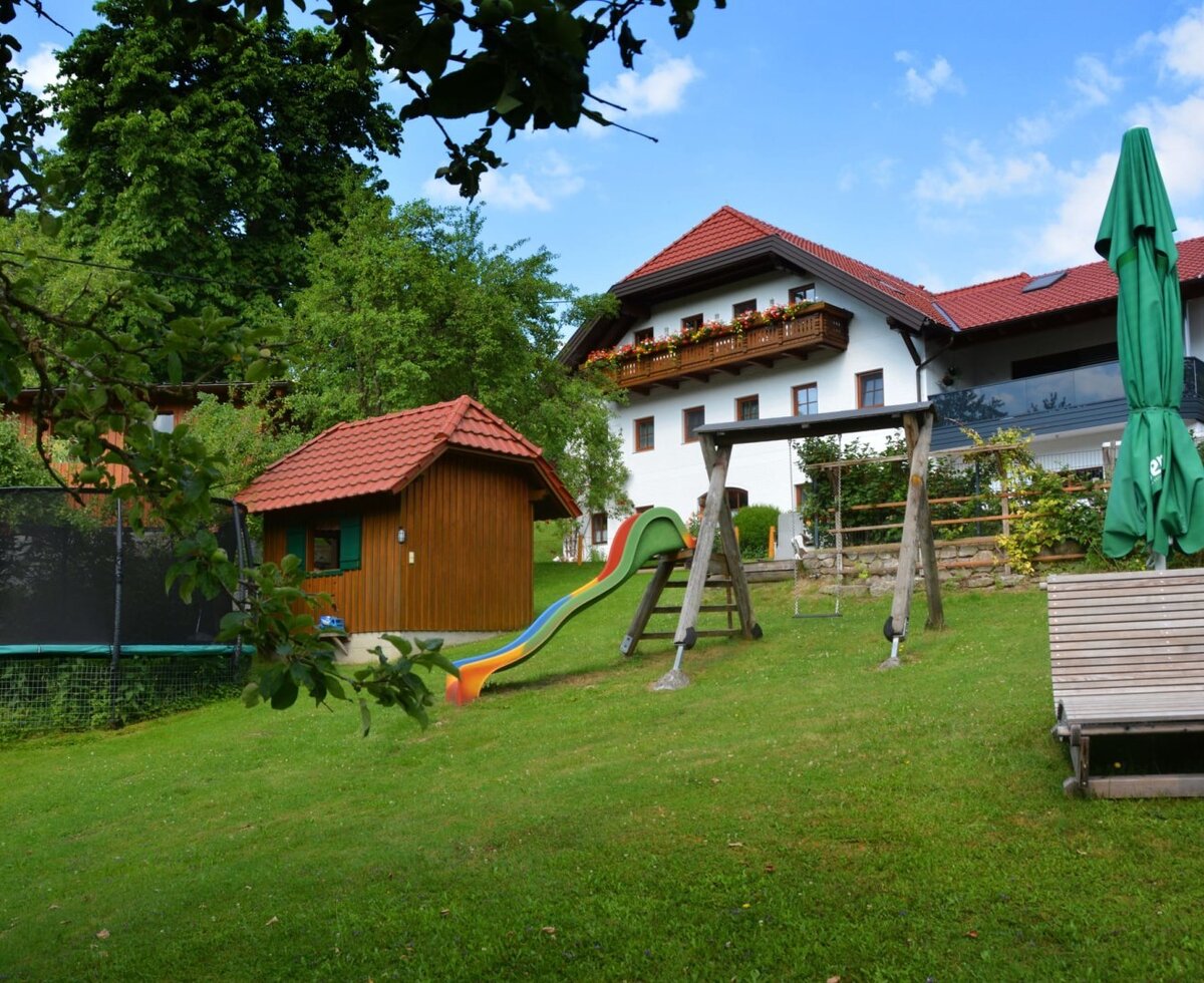 The outdoor area of the Bauernhof with a playground including a slide, swing set, and trampoline, a playhouse, and a wooden lounger on the lawn.