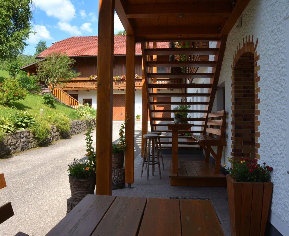 Outdoor area of the Bauernhof with a wooden staircase, a seating area with a bench and stools, and a view of another building.