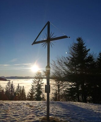 View from a summit cross on a snowy hill, overlooking a cloud inversion and the surrounding mountains.