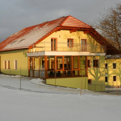 The farmhouse exterior with a light yellow facade, red tiled roof, a glass-enclosed sunroom, and a balcony, visible during winter.