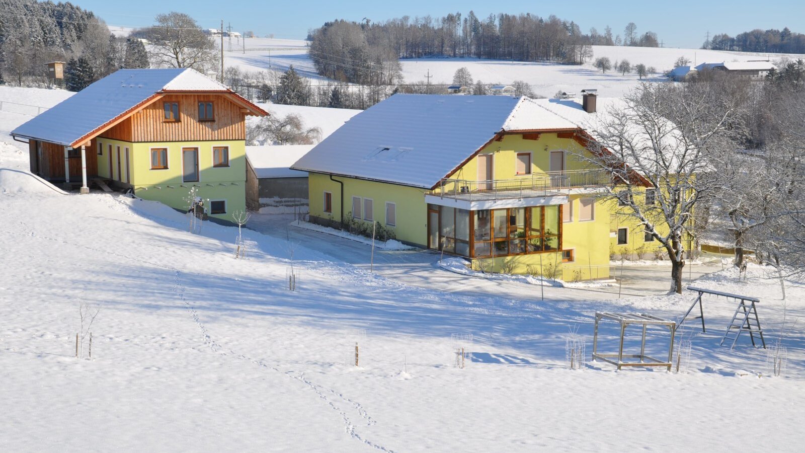 The farm house, with its two buildings, surrounded by a snowy landscape, includes a children's swing set.