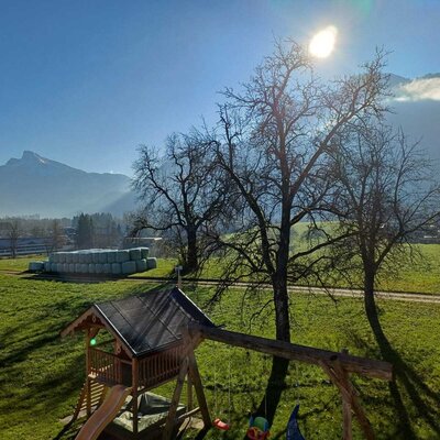 The farmhouse offers a children's playground, green fields with stacked hay bales, and views of the surrounding mountains.