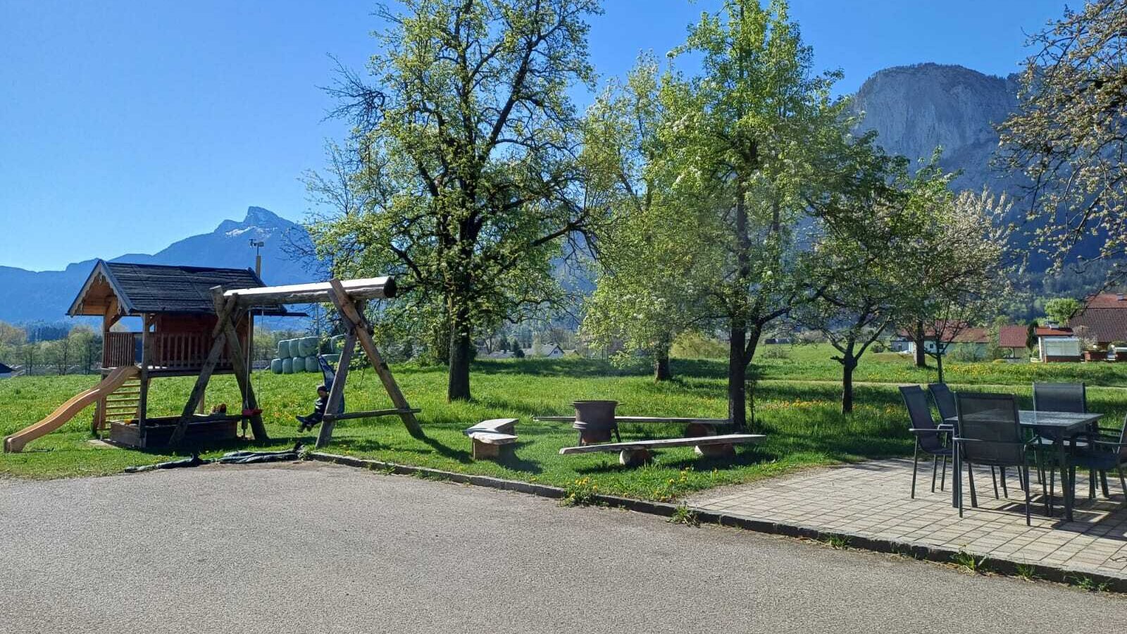 The children's playground with a playhouse and swing, and outdoor seating at the farmhouse, overlooking the surrounding meadows and mountains.
