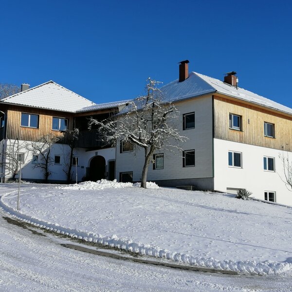 The farm house exterior with a white facade, wooden upper level, and snow-covered grounds under a clear blue sky.