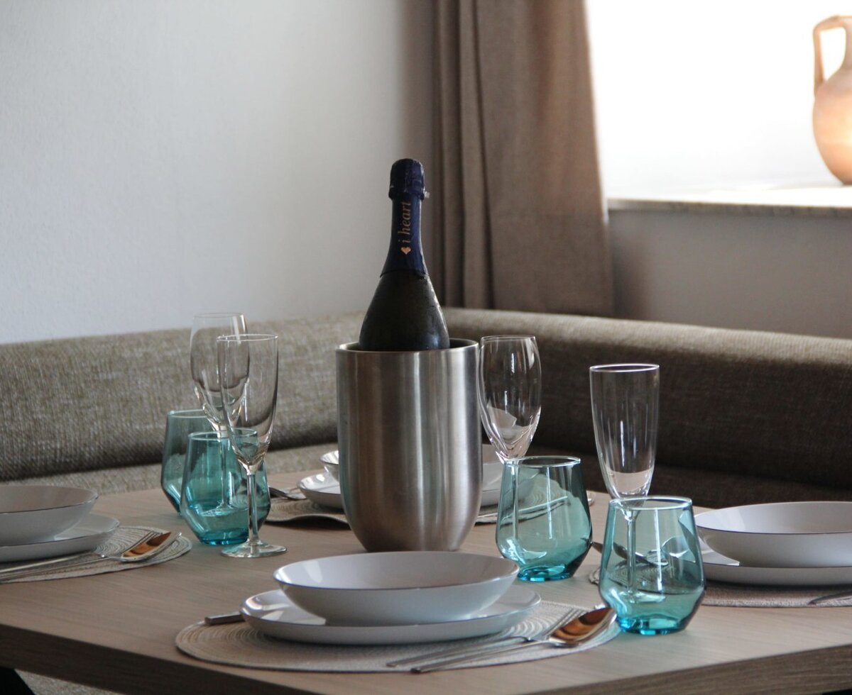 The dining table in the holiday apartment is set with white plates, blue and clear glasses, cutlery, and a bottle in an ice bucket.