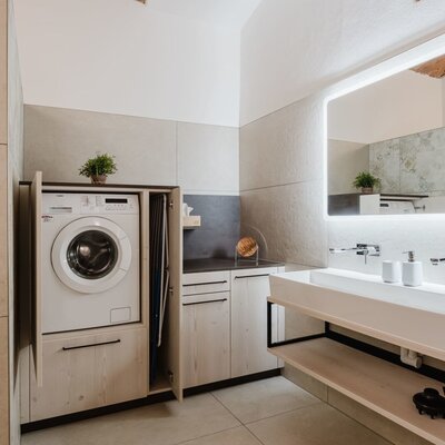Modern bathroom of the apartment with a washing machine, double sink, illuminated mirror, and shower area.
