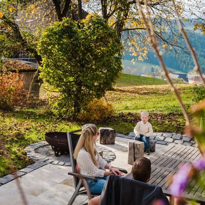 The farmhouse terrace with outdoor seating, a fire pit, and a child in the garden, offers views of the surrounding hilly landscape.
