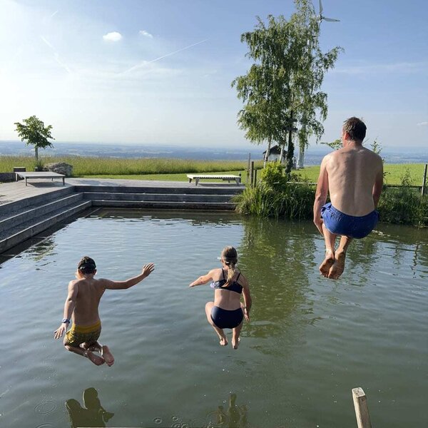 The natural swimming pond at the farmhouse, with a wooden jetty from which guests jump into the water, surrounded by landscape.