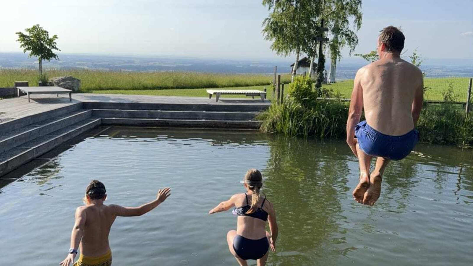 The natural swimming pond at the farmhouse, with a wooden jetty from which guests jump into the water, surrounded by landscape.