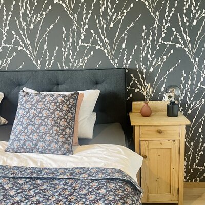 The Farm House bedroom features a bed with an upholstered headboard, patterned bedding, a wooden bedside table, and a wall with a white botanical pattern.