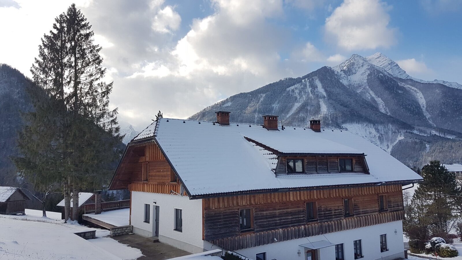The farm house with its white and wood exterior and snow-covered roof, surrounded by a snowy landscape and mountains.