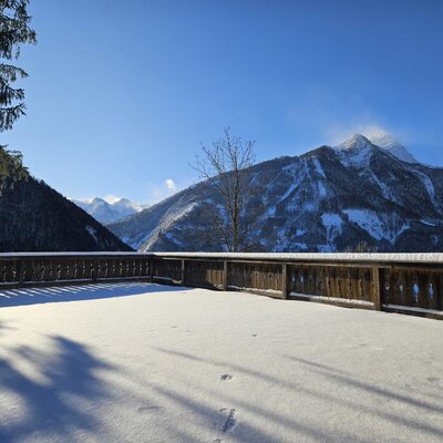 Snow-covered terrace of the farmhouse with a wooden railing and a view of the surrounding mountains and valley.