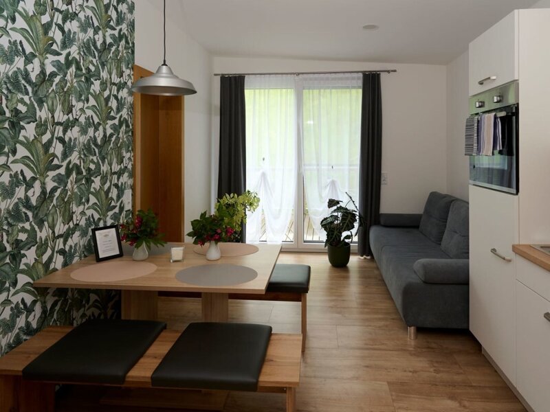 The open-plan living space of the farmhouse features a dining area with a wooden table and benches, a sofa, an integrated kitchen, and balcony access.