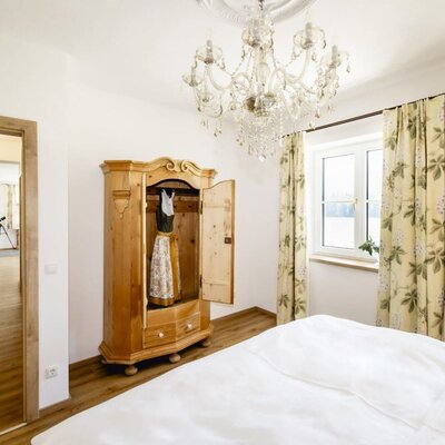 Bedroom in the apartment featuring a bed, a traditional wooden wardrobe with a dirndl, a chandelier, and windows.
