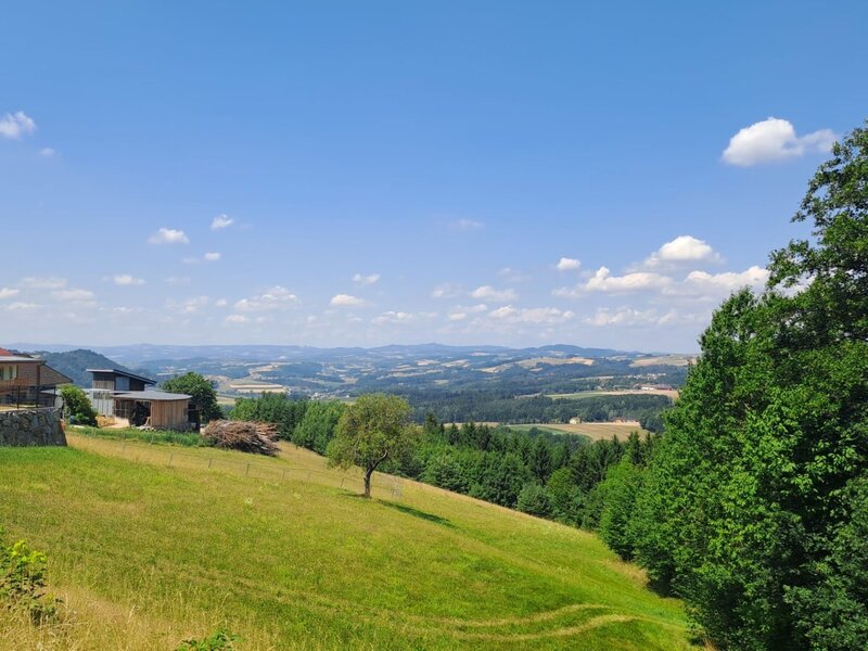 The Riederegger-Alm featuring panoramic views over the rolling green hills and forests of the Mühlviertel under a clear blue sky.