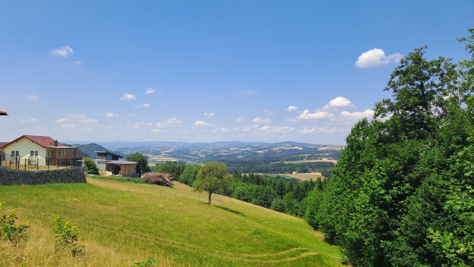 The Riederegger-Alm featuring panoramic views over the rolling green hills and forests of the Mühlviertel under a clear blue sky.
