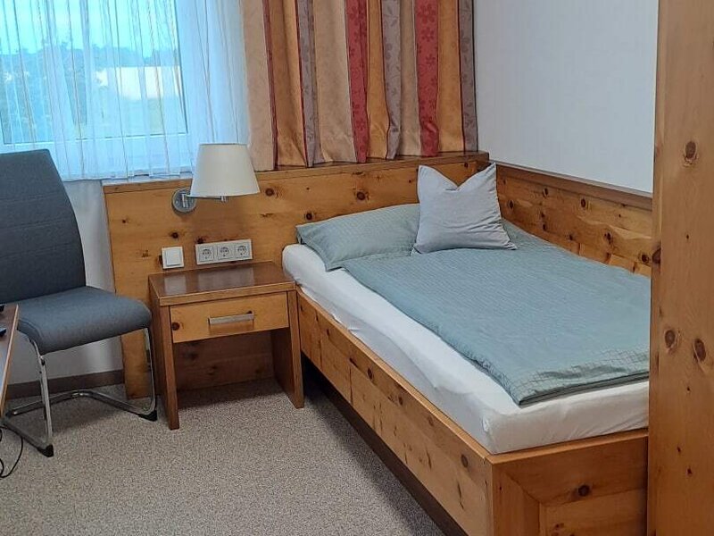 A cozy room featuring a rustic wooden single bed, a bright window, and warm wooden furnishings.