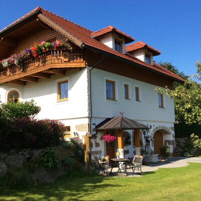The exterior of the farm house features a red-tiled roof, a wooden balcony with flowers, and an outdoor seating area on the lawn.